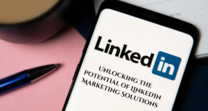 Smartphone displaying the LinkedIn logo on a screen, with the text "Unlocking the Potential of LinkedIn Marketing Solutions" overlaying the image. A pen and part of a coffee cup are visible in the background.